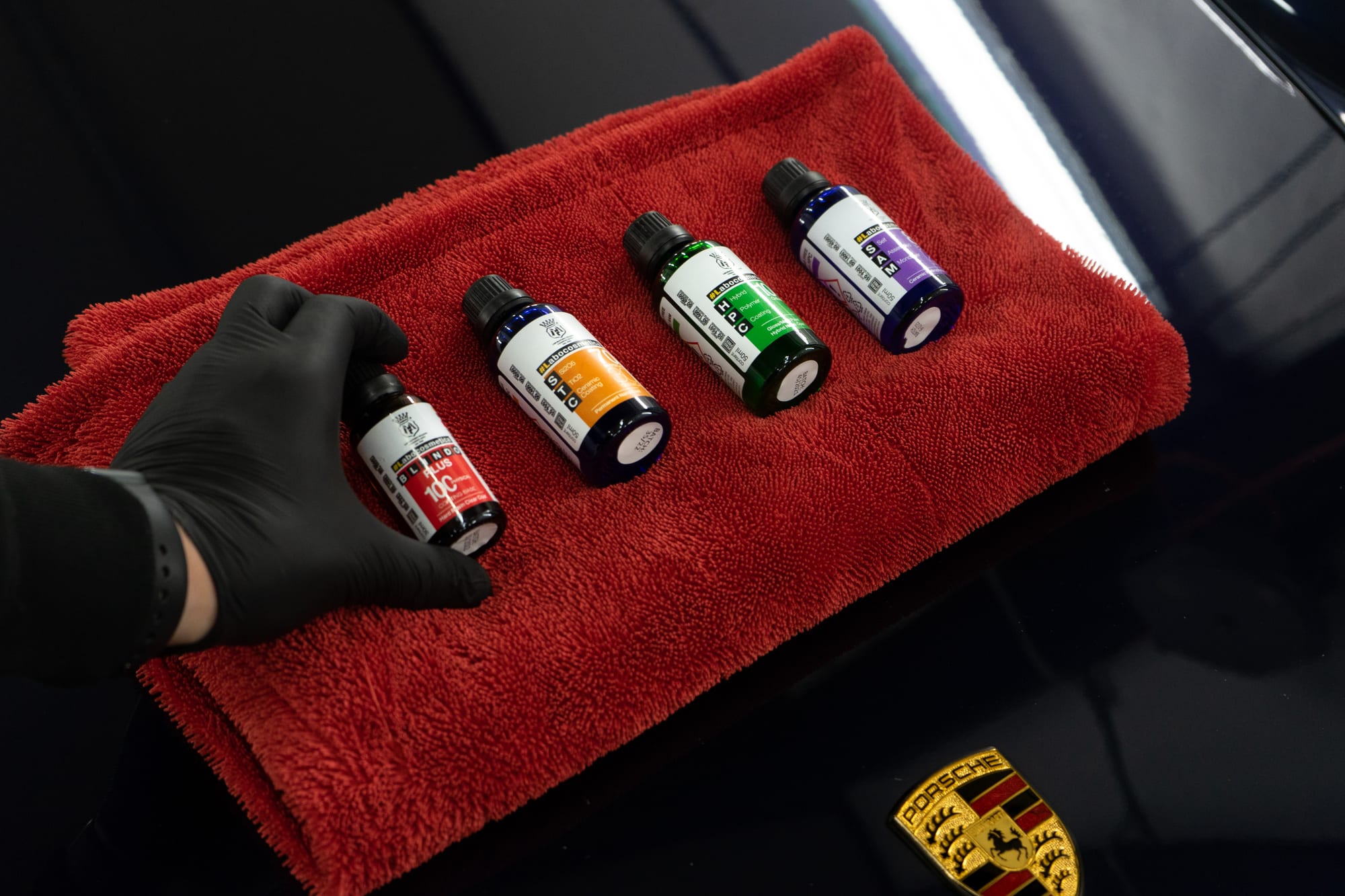 En hand i svart detailinghandske håller upp flaskor med keramisk bilvårdsbeläggning på en röd mikrofiberduk, med en Porsche-logotyp synlig i bakgrunden